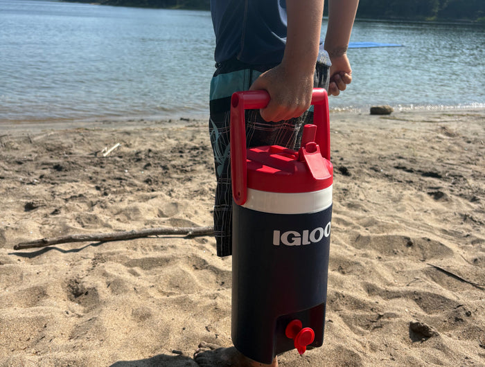 A person standing on a sandy beach by the water holds an Igloo insulated cooler with a red handle and lid, with red clogs on the sand in the foreground.